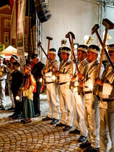 A group of people in historical uniforms stands in a square. They hold lanterns and tools in their hands while a festive atmosphere prevails. | © Foto Buchgraber