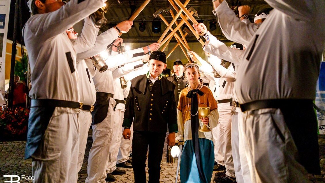 Ein festlicher Umzug mit Menschen in traditionellen Kostümen. Die beiden Protagonisten werden von einer Gruppe in weißen Kleidern mit Stöcken umrahmt. | © Foto Buchgraber
