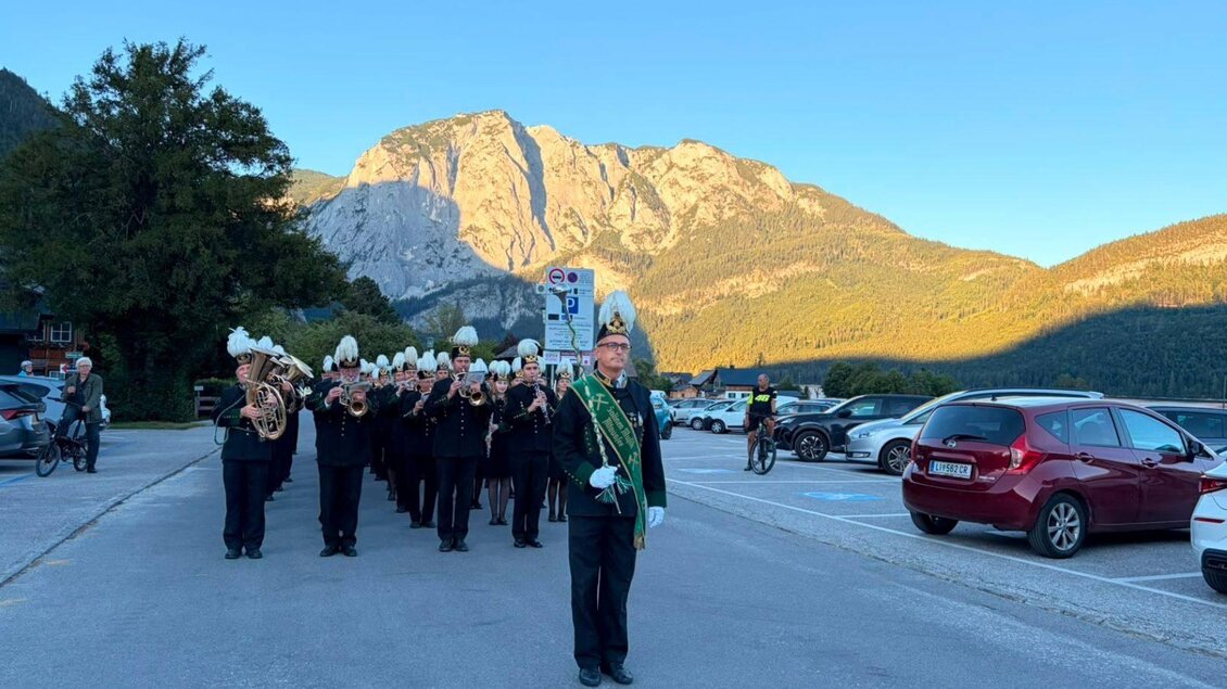 Eine Musikgruppe marschiert auf einer Straße, während die Berge im Hintergrund leuchten. Der Anführer trägt eine Schärpe und hält einen Taktstock. | © Salinenmusik Altaussee_Theresa Rastl_Robert Leu