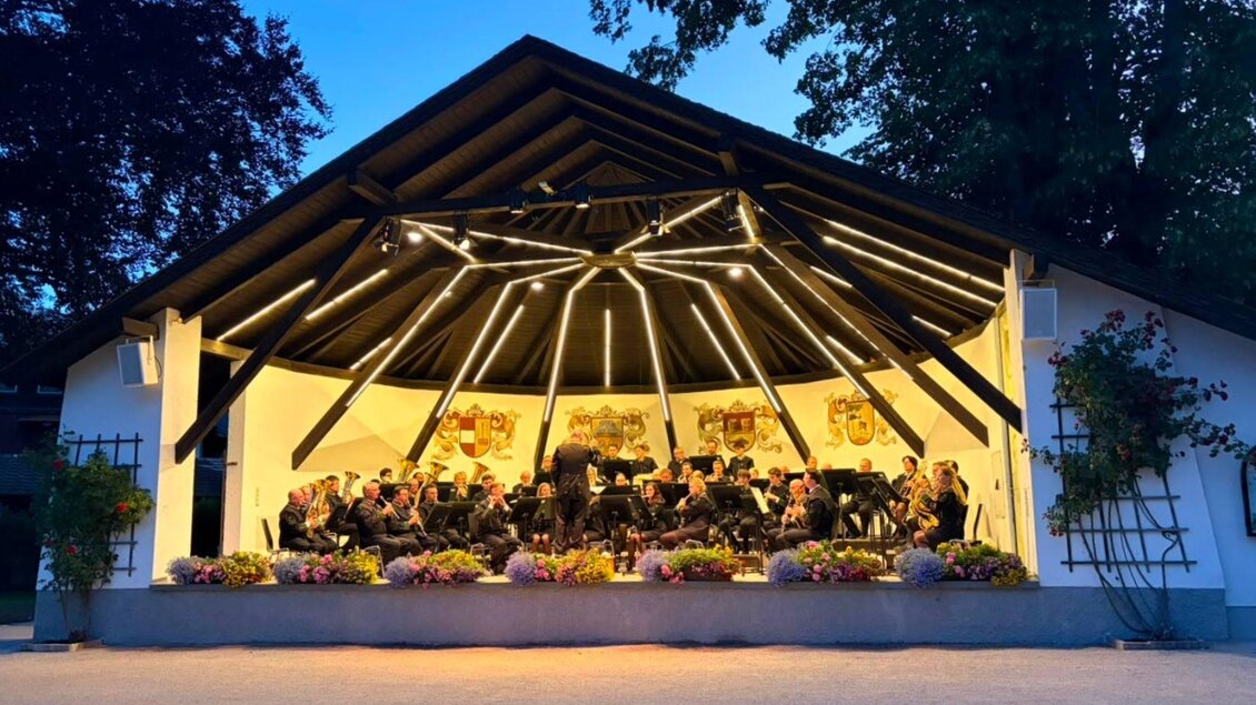 Ein Musikensemble spielt auf einer Bühne im Freien. Die Bühne ist mit bunten Blumen und Lichtern geschmückt, umgeben von Bäumen. | © Salinenmusik Altaussee_Theresa Rastl_Robert Leu