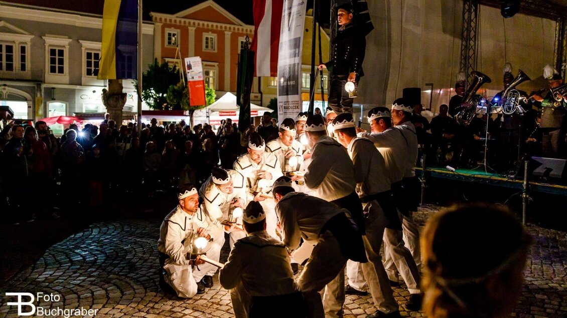 Eine Gruppe von Menschen führt eine Aufführung mit Lichtern auf einem Platz durch. Im Hintergrund sind Zuschauer und verschiedene Stände zu sehen. | © Foto Buchgraber