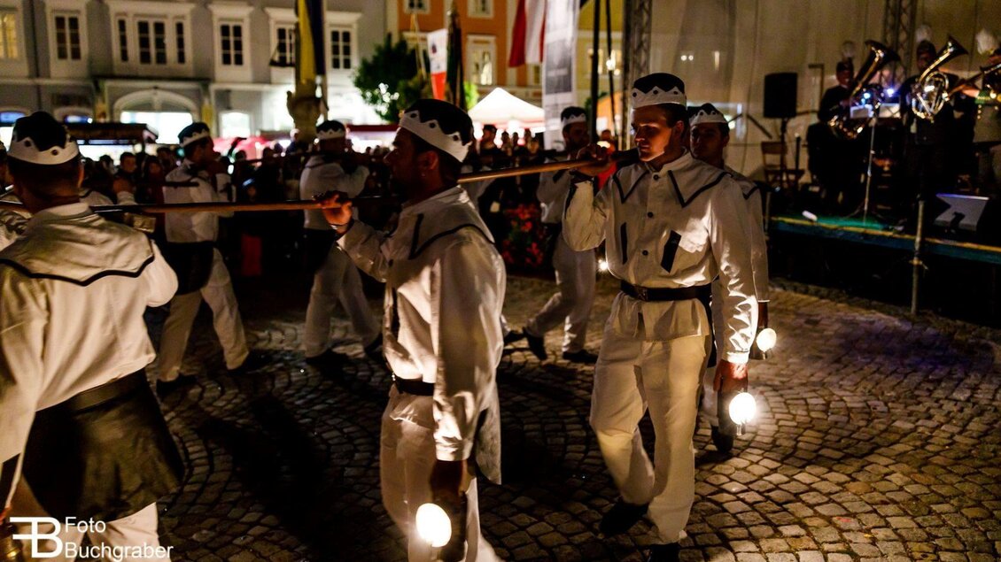 Eine Gruppe von Männern in weißen Uniformen marschiert mit Laternen in der Dunkelheit. Im Hintergrund sind Menschen und eine Band zu sehen. | © Foto Buchgraber