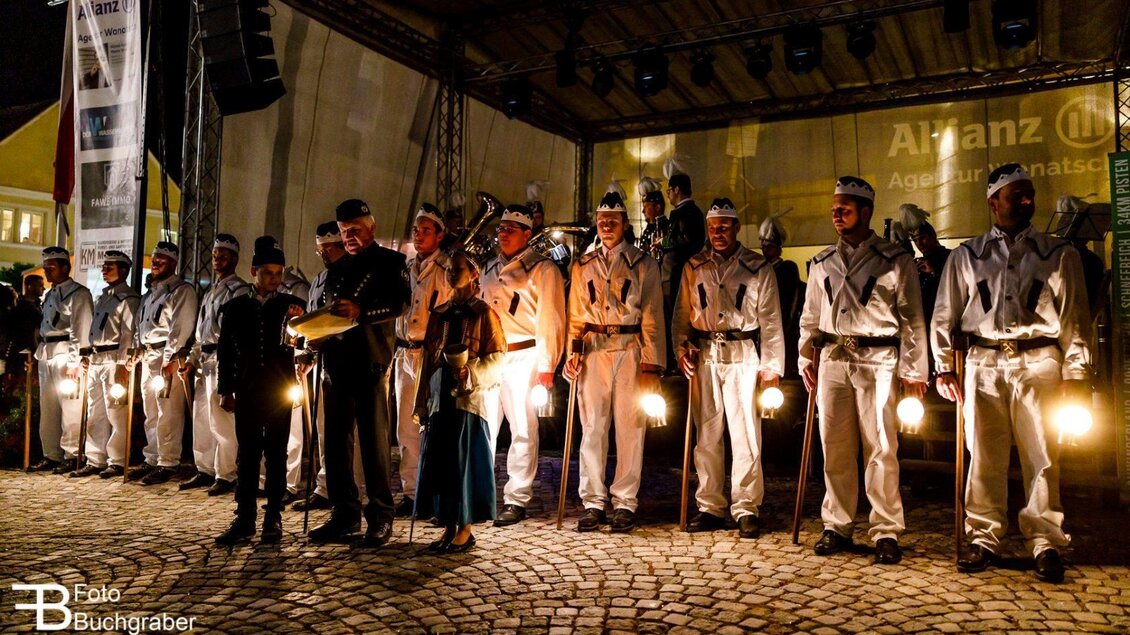 Eine Gruppe von Menschen in historischen Uniformen steht auf einer Bühne. Sie halten Laternen und sind Teil einer nächtlichen Veranstaltung. | © Foto Buchgraber