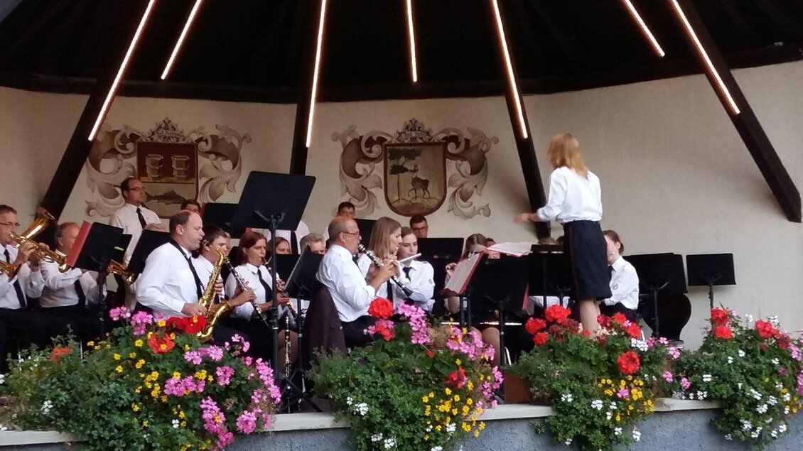 Ein Orchester spielt in einem Pavillon, während eine Dirigentin das Ensemble leitet. Bunte Blumen umrahmen die Bühne. | © Petra Kirchschlager