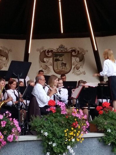 A wind orchestra plays under a roof with colorful flowers in the foreground. A conductor is leading the musicians, who are dressed in formal attire. | © Petra Kirchschlager