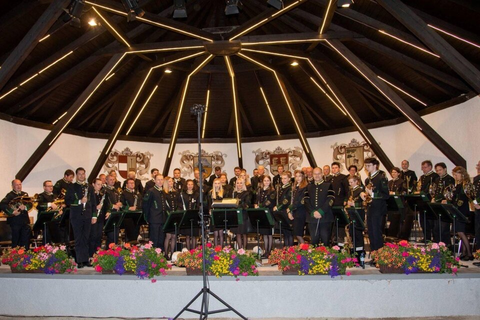 Eine Blaskapelle steht auf einer Bühne unter einem gewölbten Dach. Die Musiker tragen Uniformen und es sind bunte Blumenarrangements im Vordergrund sichtbar. | © Erich Kainzinger