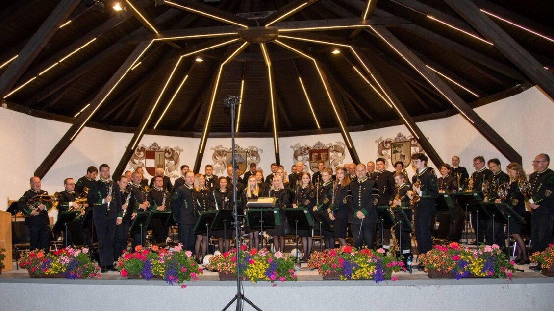 Eine Blaskapelle steht auf einer Bühne unter einem gewölbten Dach. Die Musiker tragen Uniformen und es sind bunte Blumenarrangements im Vordergrund sichtbar. | © Erich Kainzinger