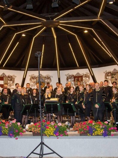 A brass band stands on a stage under a arched roof. The musicians wear uniforms and colorful flower arrangements are visible in the foreground. | © Erich Kainzinger