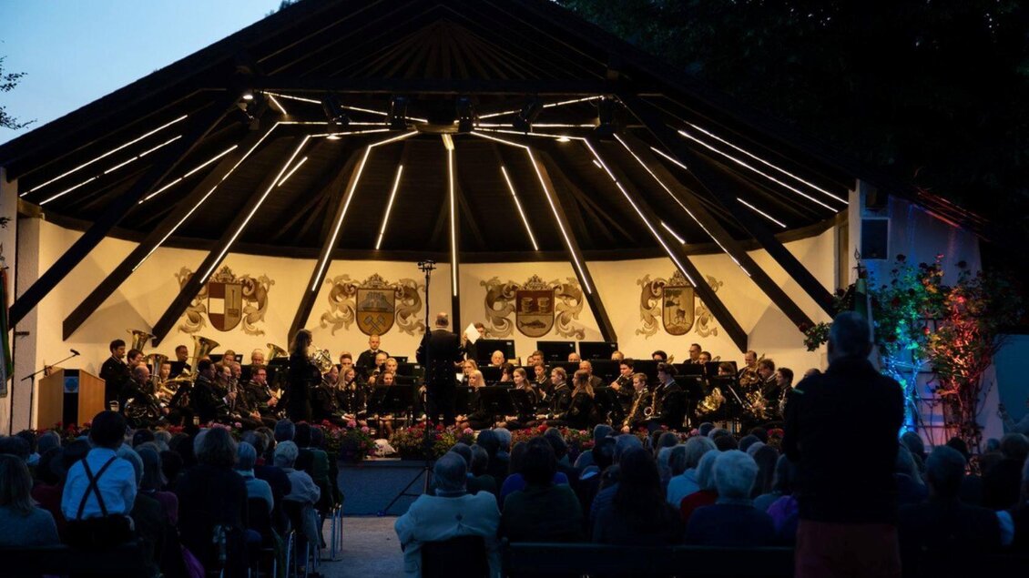Ein Orchester spielt unter einem lichtdurchfluteten Pavillon bei Abenddämmerung. Viele Zuschauer sitzen davor und genießen das Konzert. | © Erich Kainzinger