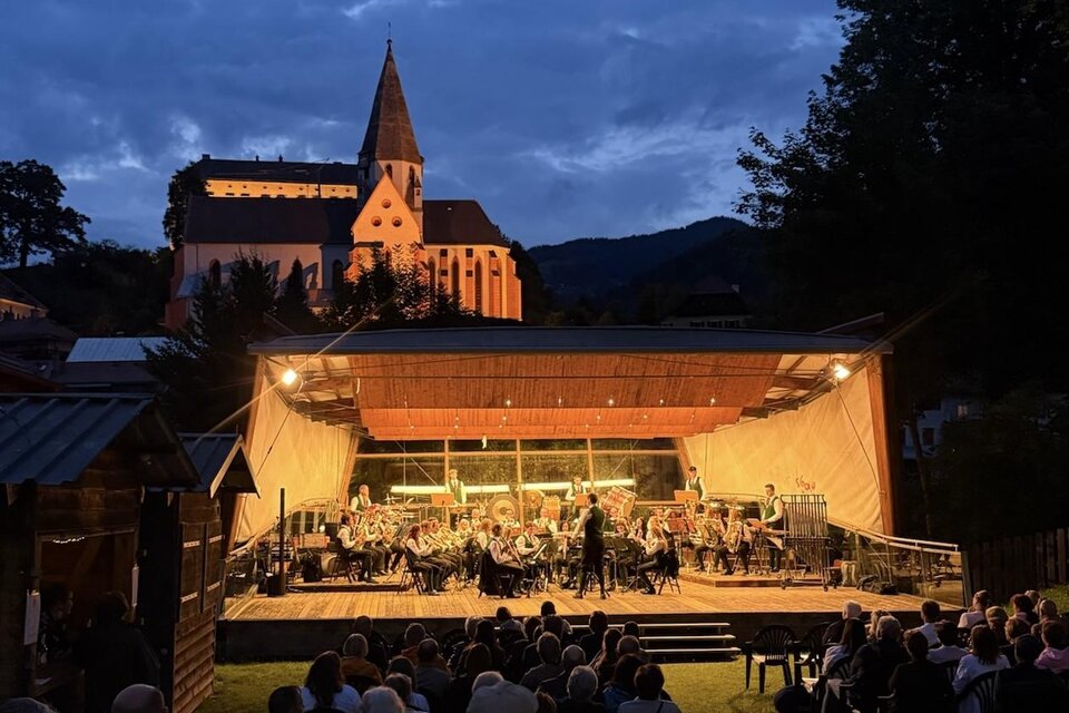 Ein Konzert im Freien bei Dämmerung mit einem großen Orchester auf der Bühne. Im Hintergrund ist eine beleuchtete Kirche und eine Zuschauermenge zu sehen.