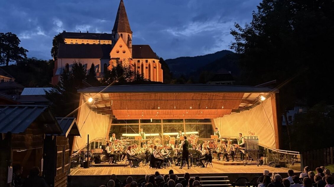Ein Konzert im Freien bei Dämmerung mit einem großen Orchester auf der Bühne. Im Hintergrund ist eine beleuchtete Kirche und eine Zuschauermenge zu sehen.