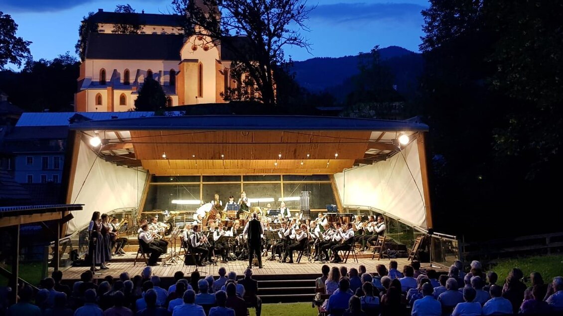 Ein Orchester spielt im Freien bei Dämmerung. Im Hintergrund ist eine Kirche und Bäume zu sehen.