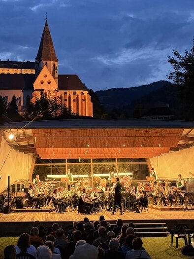 Ein Orchestervorführung im Freien bei Dämmerung, mit einer schönen Kirche im Hintergrund. Das Publikum genießt die Musik auf einer Bühne mit angemessener Beleuchtung.