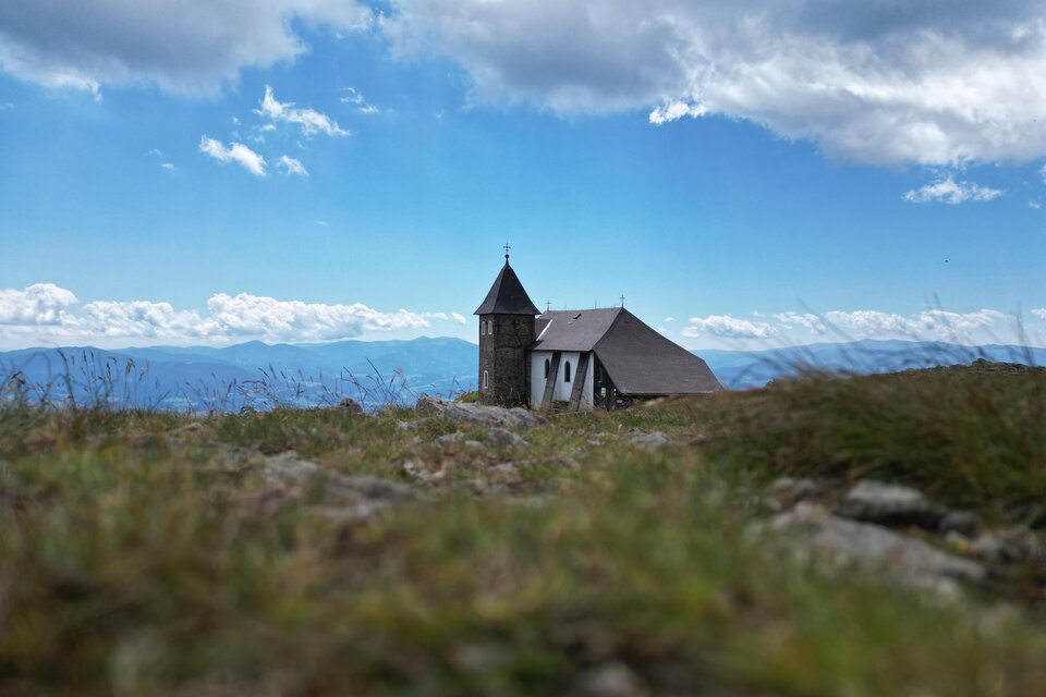 Eine kleine Kirche auf einem grünen Hügel. Im Hintergrund erstreckt sich ein klarer blauer Himmel mit einigen Wolken. | © Isabella Painhapp