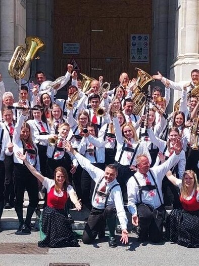Eine große Musikgruppe steht fröhlich vor einem Gebäude. Die Mitglieder tragen traditionelle Trachten und halten ihre Instrumente in die Luft. | © TV Murau
