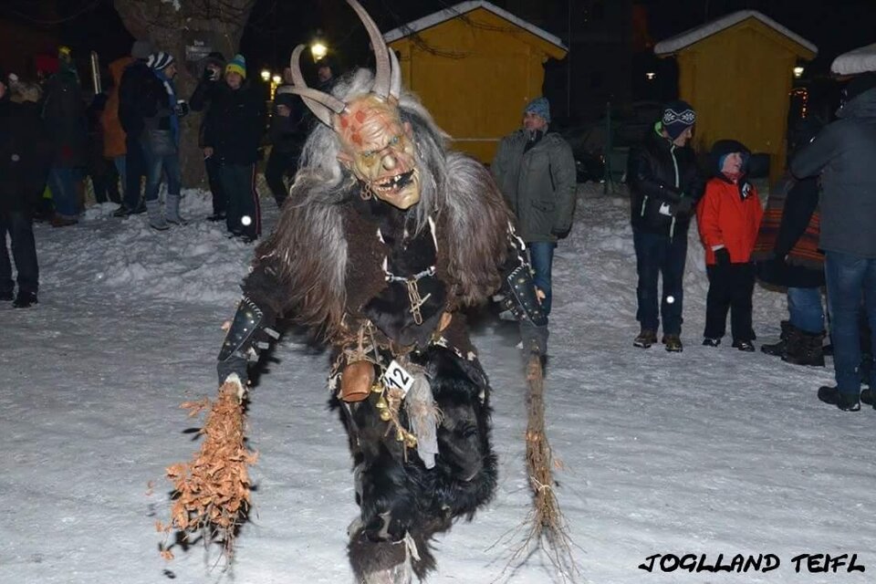 Ein gruselig verkleideter Dämon mit Hörnern und einem wilden Kostüm steht im Schnee. Um ihn herum sind Menschen in warmer Kleidung zu sehen. | © Joglland Teifl