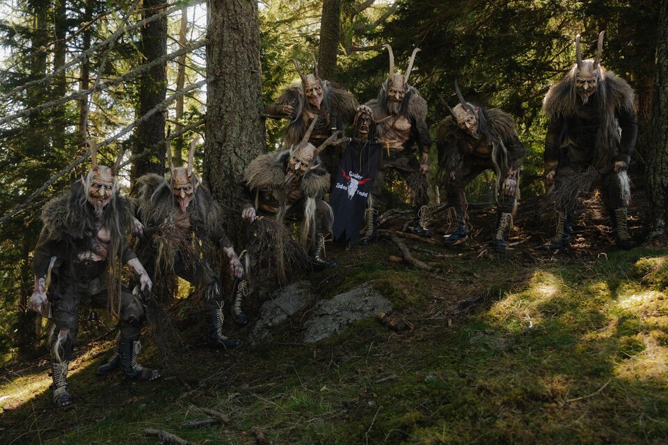 Ein festlich gekleideter Charakter mit langen Hörnern und einem furchterregenden Gesichtsausdruck. Im Hintergrund sind fröhliche Menschen zu sehen, die die Szene beobachten. | © Gaaler Zinkenteufel