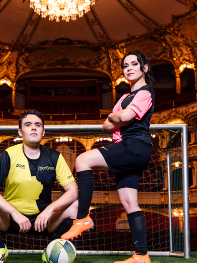 Zwei Fußballspieler posieren vor einem Tor in einem eleganten Theater. Der Mann kniet, während die Frau selbstbewusst steht und den Ball hält. | © Oper Graz Ingo Pertramer