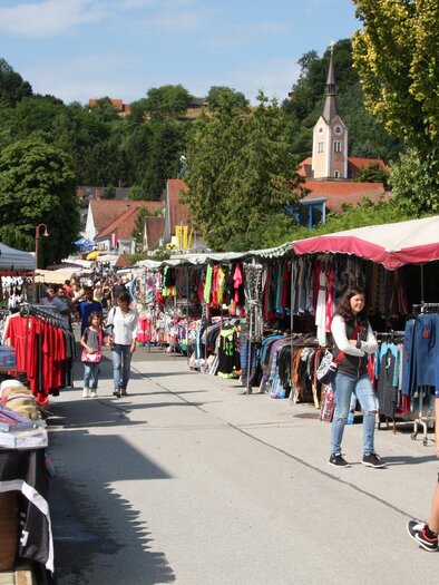 Ein geschäftiger Markt mit Ständen, die Kleidung und Accessoires verkaufen. Menschen schlendern entlang der Marktstraße unter blauem Himmel. | © Stadtgemeinde Fehring