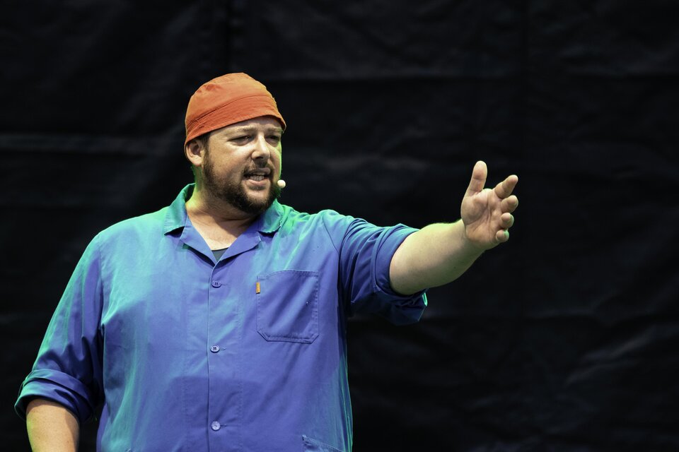 Ein Mann steht auf einer Bühne und spricht leidenschaftlich. Er trägt ein blaues Hemd und eine orangefarbene Mütze. | © C. Drobny