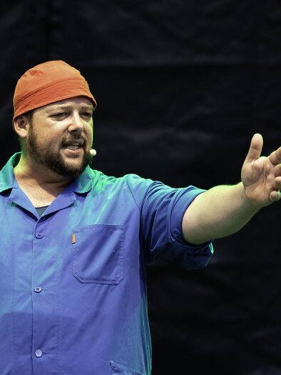 Ein Mann steht auf einer Bühne und spricht leidenschaftlich. Er trägt ein blaues Hemd und eine orangefarbene Mütze. | © C. Drobny