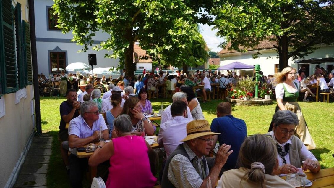 Ein belebter Garten mit zahlreichen Gästen, die an Tischen sitzen und essen. Im Hintergrund sind Bäume und Festzelte zu sehen. | © Peter Schandor