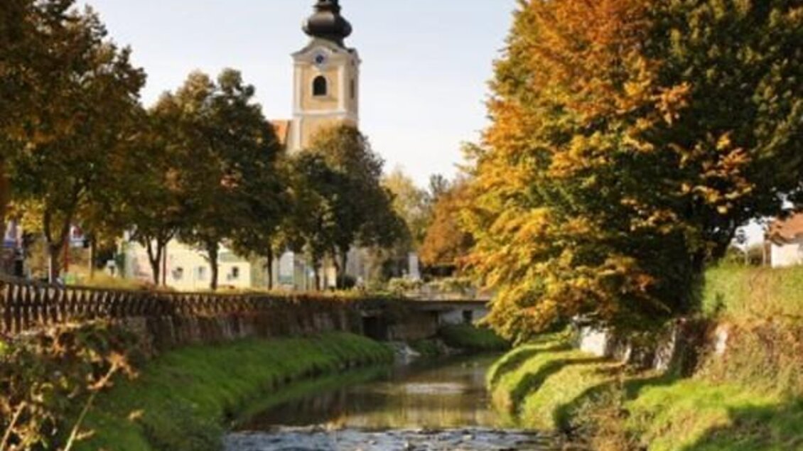 Ein ruhiger Fluss fließt durch eine malerische Landschaft mit bunten Bäumen im Herbst. Im Hintergrund ist eine Kirche mit einem einzigartigen Turm zu sehen. | © Bergmann