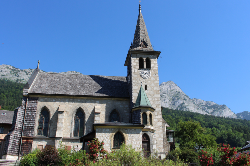 Eine malerische Kirche mit einem hohen Turm, umgeben von grünen Bäumen und Bergen. Der klare blaue Himmel ergänzt die friedliche Landschaft. | © Viola Lechner