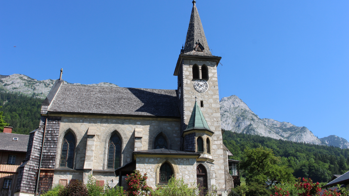 Eine malerische Kirche mit einem hohen Turm, umgeben von grünen Bäumen und Bergen. Der klare blaue Himmel ergänzt die friedliche Landschaft. | © Viola Lechner