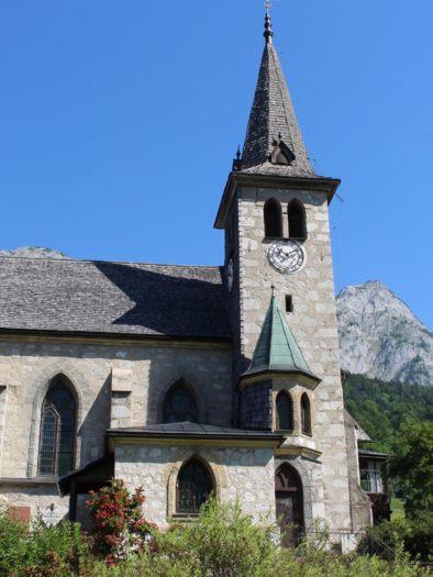 A picturesque church with a tall tower, surrounded by green trees and mountains. The clear blue sky complements the peaceful landscape. | © Viola Lechner