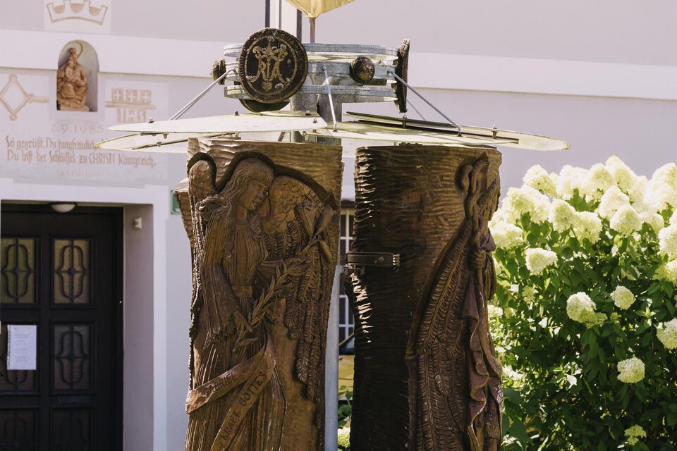 Eine kunstvolle Skulptur steht in einem grünen Garten. Im Hintergrund sind einen Gebäudefassade und blühende Pflanzen zu sehen.