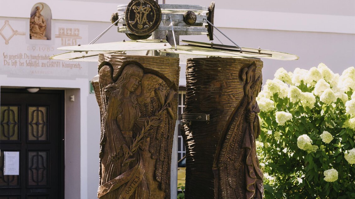 Eine kunstvolle Skulptur steht in einem grünen Garten. Im Hintergrund sind einen Gebäudefassade und blühende Pflanzen zu sehen.