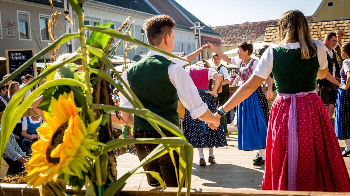 Eine festliche Tanzveranstaltung im Freien mit Menschen in traditionellen Trachten. Im Vordergrund sind Sonnenblumen und eine lebendige Atmosphäre zu sehen. | © @stefanfriesinger