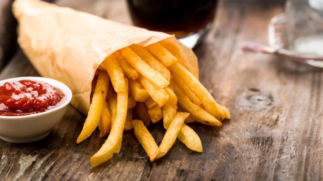 Eine Tüte frischer Pommes frites liegt auf einem Holztisch. Neben der Tüte befindet sich eine kleine Schale mit Ketchup.