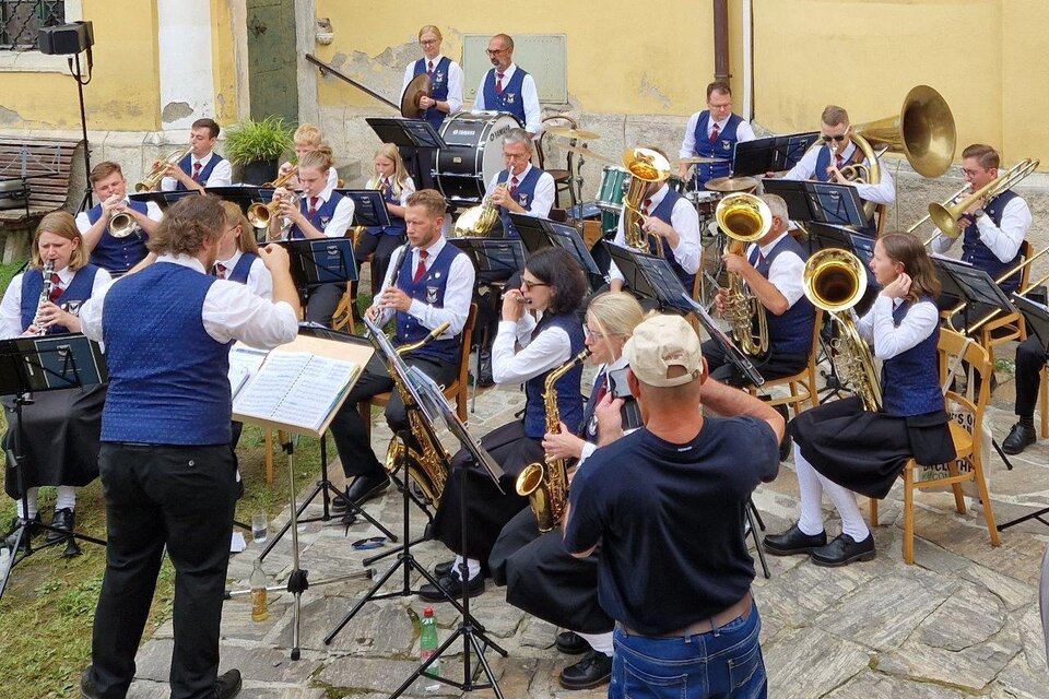 Eine Blaskapelle spielt im Freien, angeführt von einem Dirigenten. Die Musiker tragen traditionelle Kostüme und es sind verschiedene Instrumente zu sehen. | © Konrad Edegger