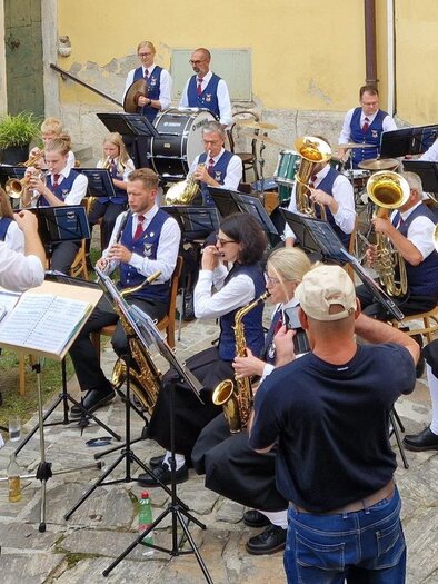Eine Blaskapelle spielt im Freien, angeführt von einem Dirigenten. Die Musiker tragen traditionelle Kostüme und es sind verschiedene Instrumente zu sehen. | © Konrad Edegger