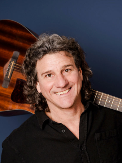 A man is smiling and holding an acoustic guitar over his shoulder. The background is dark blue and highlights the guitar. | © Pressefoto_Miriam Mehlman