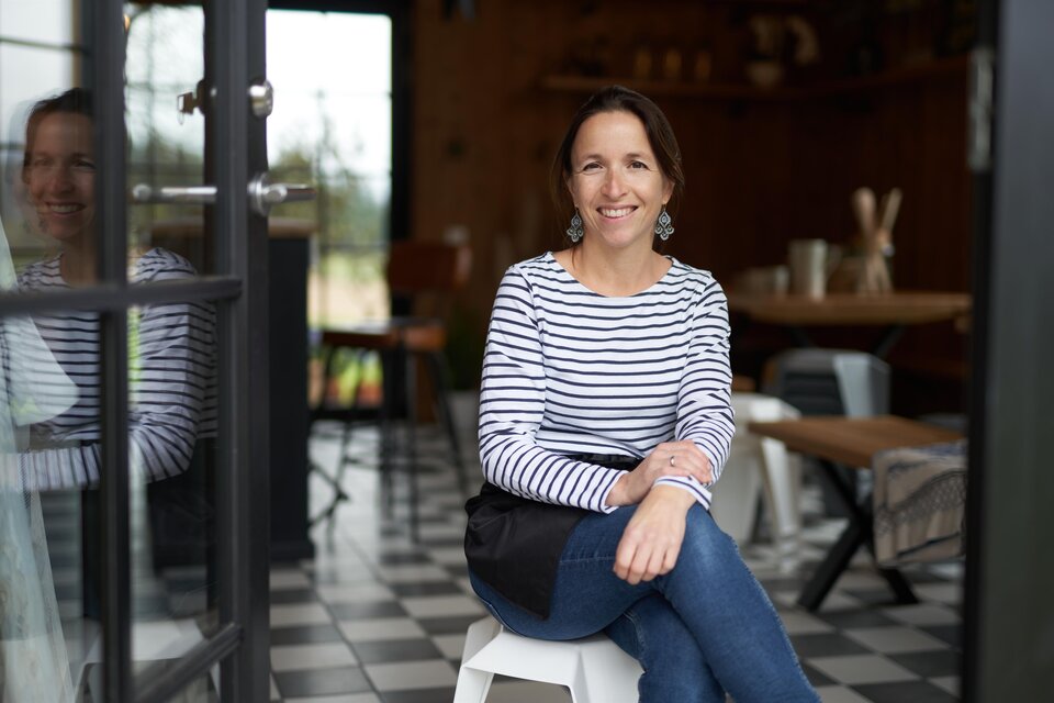 Eine Frau sitzt lächelnd auf einem Hocker in einem gemütlichen Café. Der Raum ist stilvoll eingerichtet mit einem einladenden Ambiente. | © Markus Lang Bichl