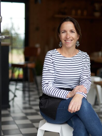 Eine Frau sitzt lächelnd auf einem Hocker in einem gemütlichen Café. Der Raum ist stilvoll eingerichtet mit einem einladenden Ambiente. | © Markus Lang Bichl