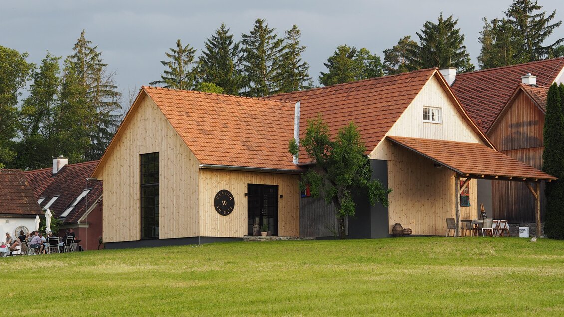 Ein modernes Haus mit einem Holzverkleidung und einem Satteldach liegt in einer grünen Wiese. Im Hintergrund sind Bäume und weitere Gebäude zu sehen. | © Karin Heschl-Polzhofer