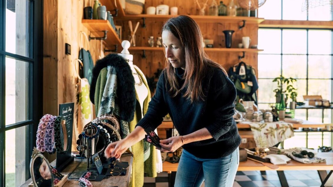 Eine Frau arrangiert Schmuck in einem gemütlichen Laden. Im Hintergrund sind Kochgeschirr und eine grüne Jacke zu sehen. | © Karin Heschl-Polzhofer