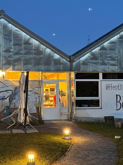 Ein modernes Gebäude mit großen Fenstern und einem bunten Wandbild einer Eule. Vor der Tür stehen zwei weiße Tische und Stühle in einem einladenden Garten. | © Bücherei Stubenberg am See