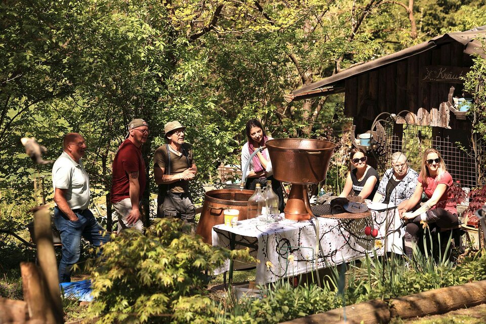 Eine Gruppe von Menschen sitzt und steht in einem Gartenbereich neben einem Tisch mit verschiedenen Utensilien. Im Hintergrund sind Bäume und ein kleines Gebäude zu sehen. | © Mike & The Muse