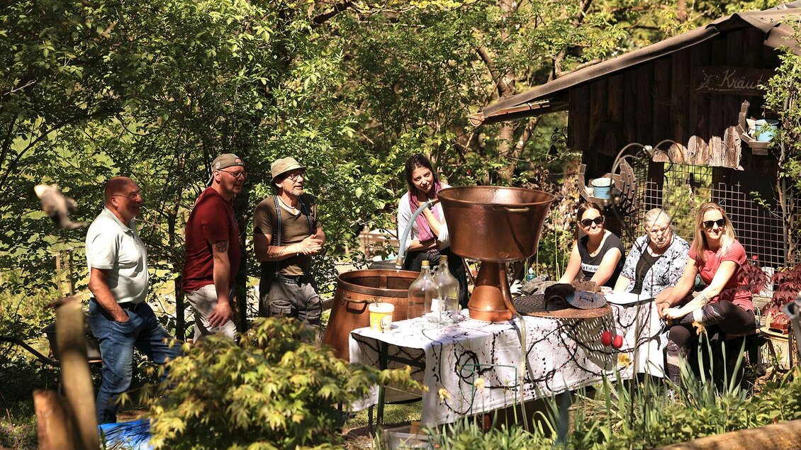 Eine Gruppe von Menschen sitzt und steht in einem Gartenbereich neben einem Tisch mit verschiedenen Utensilien. Im Hintergrund sind Bäume und ein kleines Gebäude zu sehen. | © Mike & The Muse