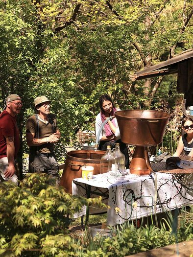 A group of people is sitting and standing in a garden area next to a table with various utensils. In the background, trees and a small building can be seen. | © Mike & The Muse