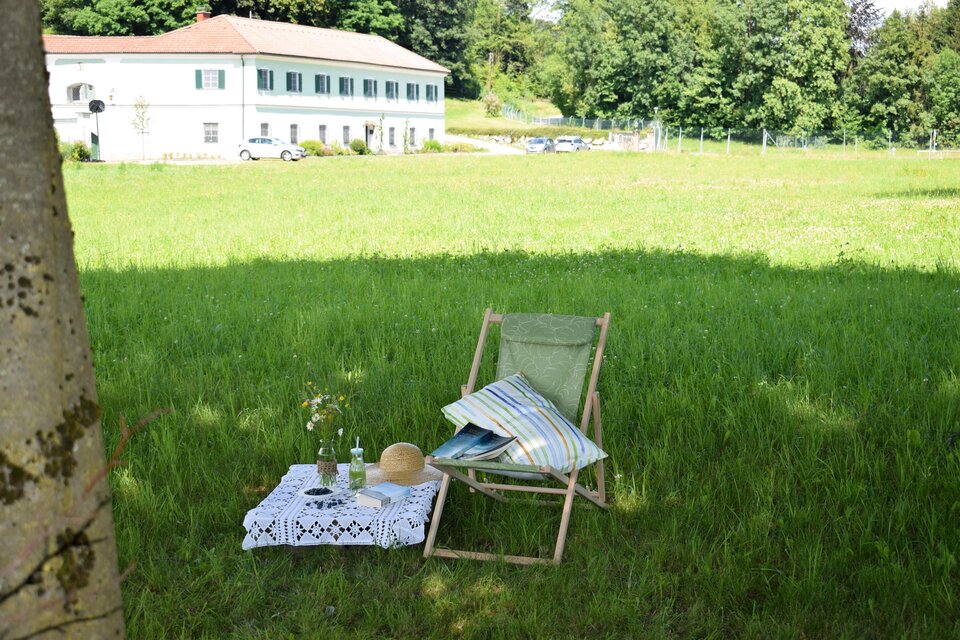 Eine gemütliche Sitzecke auf einer grünen Wiese mit einem Stuhl und einem kleinen Tisch. Im Hintergrund ist ein Gebäude und Bäume zu sehen. | © Gabriele Paar