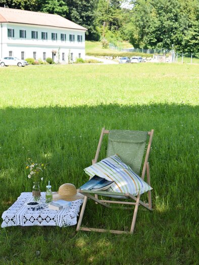 Eine gemütliche Sitzecke auf einer grünen Wiese mit einem Stuhl und einem kleinen Tisch. Im Hintergrund ist ein Gebäude und Bäume zu sehen. | © Gabriele Paar