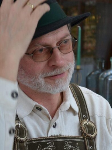 A man with glasses and a traditional hat is sitting in a cozy restaurant. He is wearing leather pants and has a friendly face. | © Familie Michelitsch