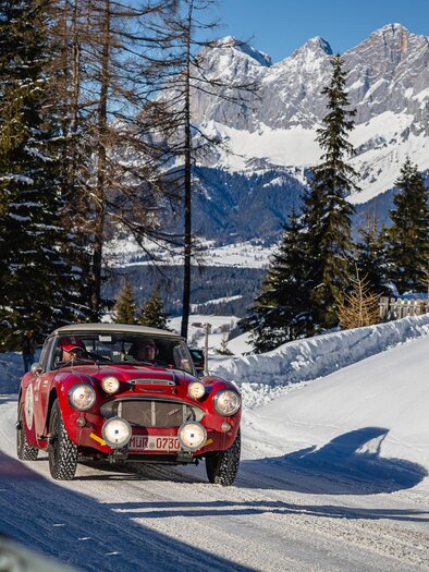 A classic car drives along a snowy road in the mountains. In the background, tall mountains and coniferous trees are visible. | © Steering Media