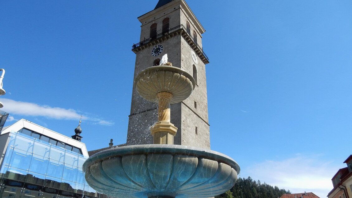 Ein schöner Brunnen mit einer alten Turmuhr im Hintergrund. Der Himmel ist klar und blau, und der Platz ist sonnig. | © Sternenturm Judenburg